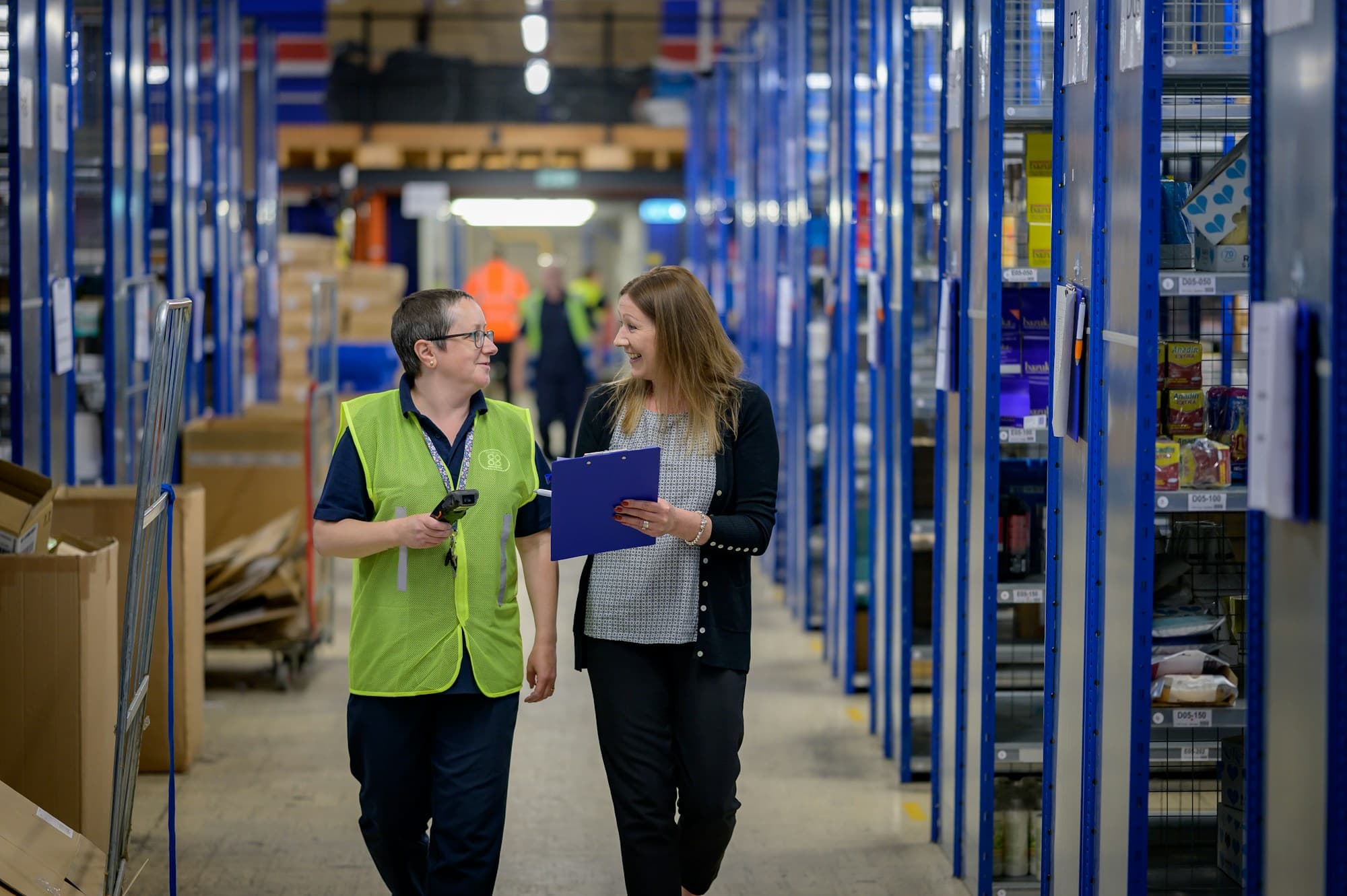 Two people in hi-vis walking through a warehouse aisle
